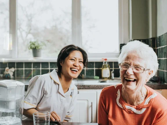 An elderly woman laughs beside a friendly young care assistant